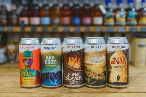 Collection of Buxton beer cans on a wooden surface with a blurred background of more bottles.