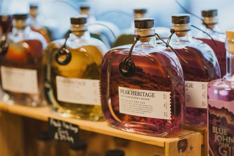 Row of bottles with labels on a wooden shelf, featuring 'Peak Heritage' and 'Blue John Pink Gin'.