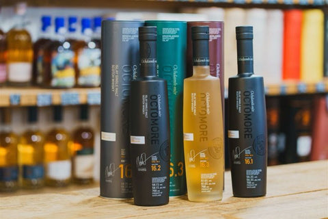 Four bottles of Octomore whisky with packaging on a wooden surface, blurred shelves in the background.